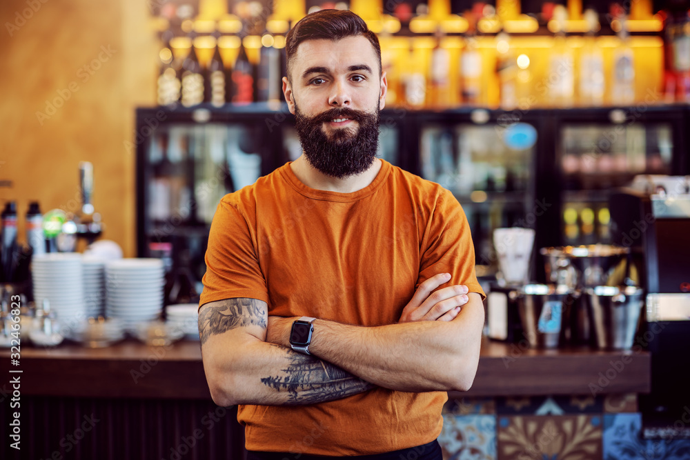 Young handsome bearded tattooed cafe owner standing with arms crossed and looking at camera. Young handsome bearded tattooed cafe owner standing with arms crossed and looking at camera.