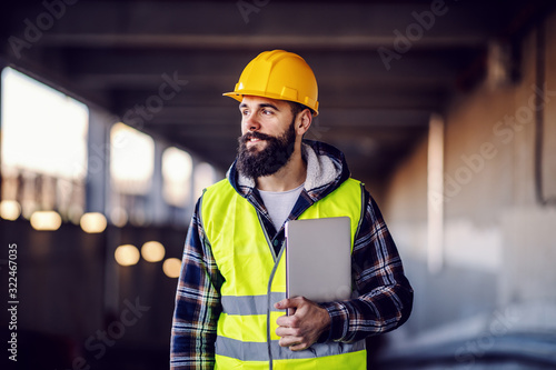 Fototapeta Portrait of highly motivated caucasian hardworking smiling bearded supervisor with helmet on head in vest and with laptop in hands posing on construction site