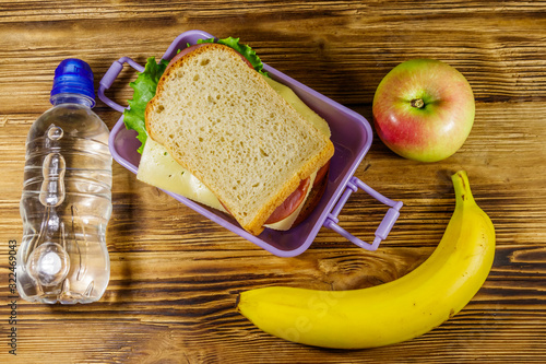 Lunch box with sandwiches, bottle of water, banana and apple on a wooden table. Top view