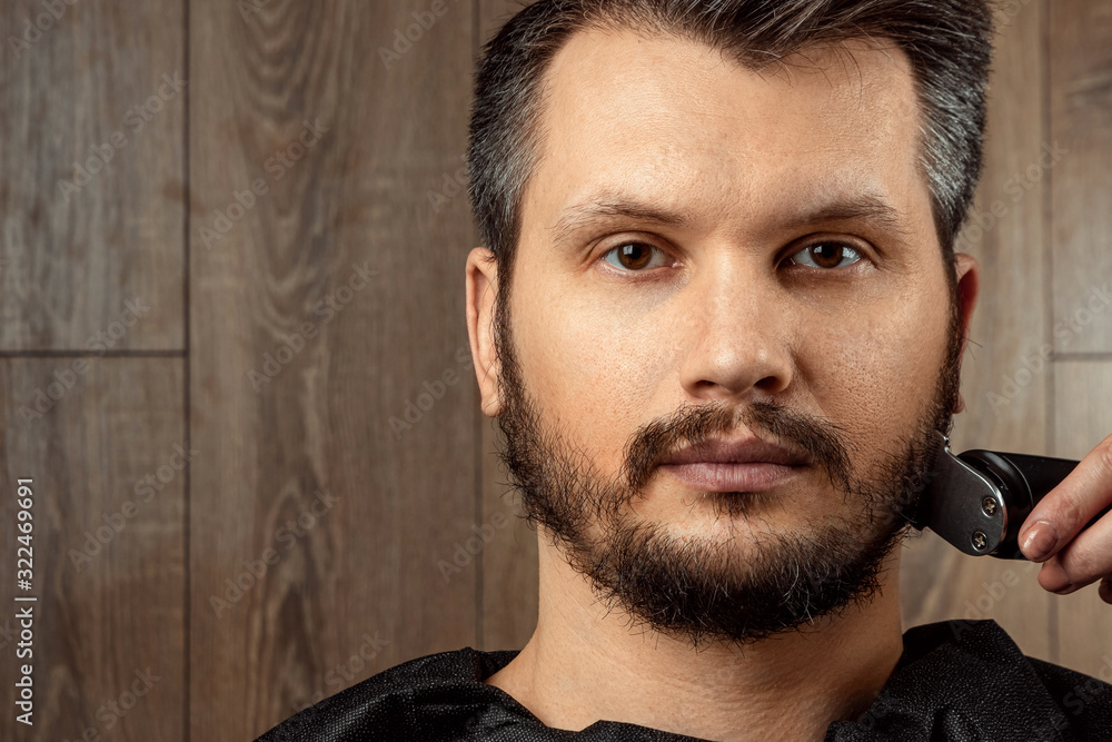 Obraz premium The master makes a beard haircut to the client with an electric machine, trimmer, close-up. The process of shaving a beard at the hairdresser. Body care, lifestyle, metrosexual.