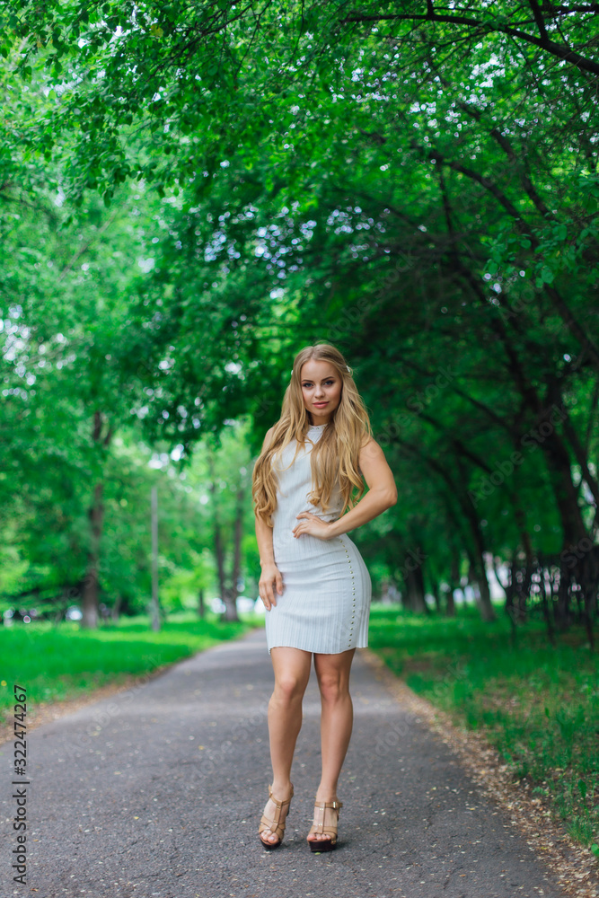 Portrait of a charming blond woman wearing beautiful white dress standing on the road under trees. Portrait of a charming blond woman wearing beautiful white dress standing on the road under trees.