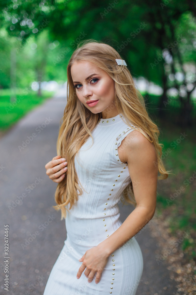 Portrait of a charming blond woman wearing beautiful white dress standing on the road under trees. Portrait of a charming blond woman wearing beautiful white dress standing on the road under trees.