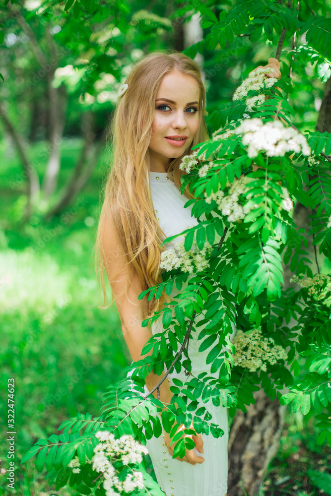 Portrait of a charming blond woman wearing beautiful white dress standing next to rowan tree with white flowers. Portrait of a charming blond woman wearing beautiful white dress standing next to rowan tree with white flowers.