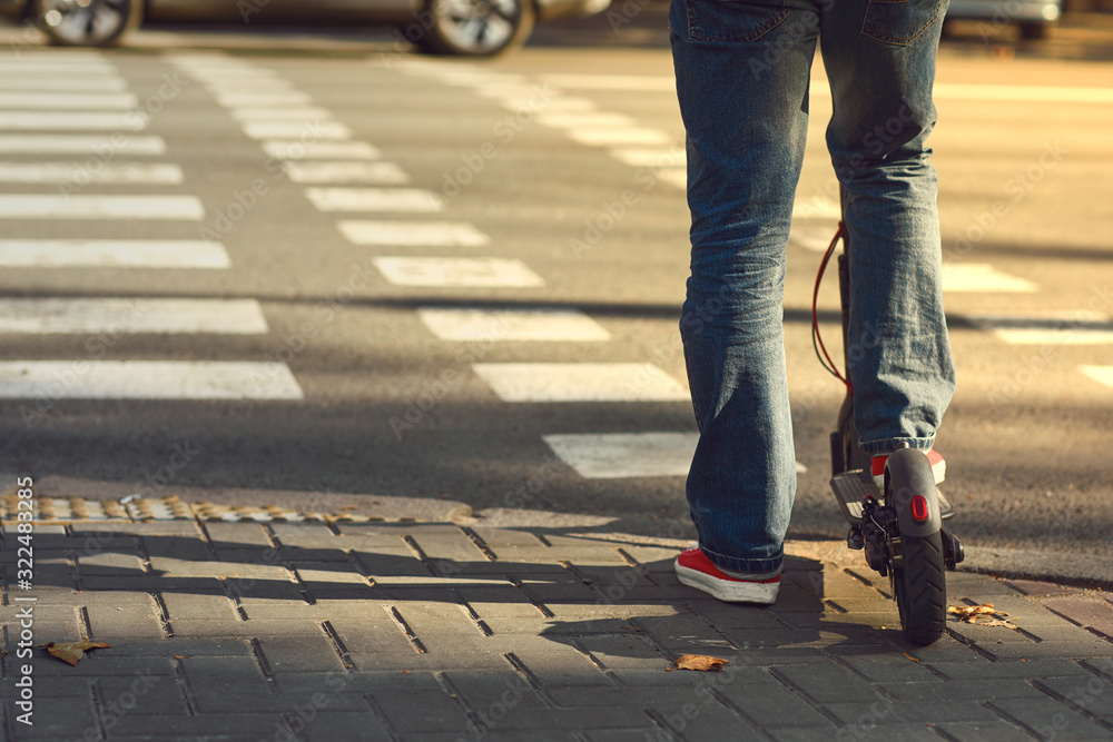 Legs of a man in jeans and sneakers on an electric scooter at a crosswalk on a city street