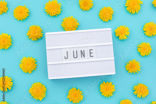 Text June on light box and yellow dandelions on blue background. Concept hello summer. Top view Flat lay.