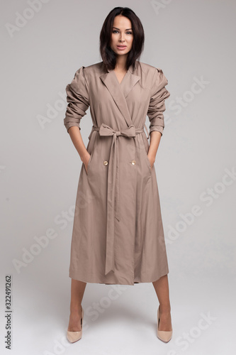 Full length of beautiful brunette young woman wearing long trench coat against grey background. Fashion and style concept