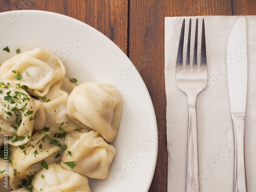Portion of Russian classic big size dumplings on a white plate with tomato ketchup and herbs. Dark wood table surface. Fork and knife on a white napkin.