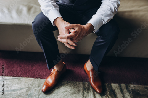 man in a white shirt, suit and brown leather shoes sitting on a sofa clasped hands