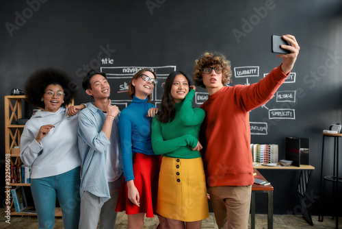 Selfie time. Group of happy multicultural people in casual wear making selfie on the phone while standing against blackboard in the modern office. Young cheerful designers looking into the camera