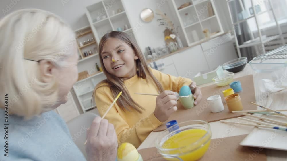 Zoom in shot of happy grandmother putting gouache on nose of surprised little girl and laughing together with her while decorating Easter eggs together at kitchen table Zoom in shot of happy grandmother putting gouache on nose of surprised little girl and laughing together with her while decorating Easter eggs together at kitchen table