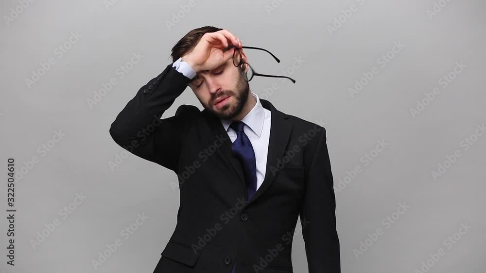 Tired bearded young business man in classic black suit take off glasses isolated on grey background studio. Achievement career wealth business concept. Mock up copy space. Rub eyes, put hand on nose Tired bearded young business man in classic black suit take off glasses isolated on grey background studio. Achievement career wealth business concept. Mock up copy space. Rub eyes, put hand on nose