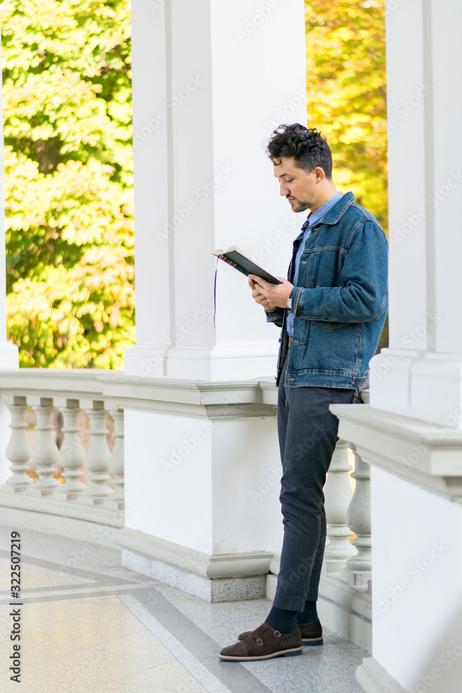 Handsome young man reading a book in a park. Portrait of a young man with a denim jacket and blue shirt reading a book outside. A guy reading a book in a park