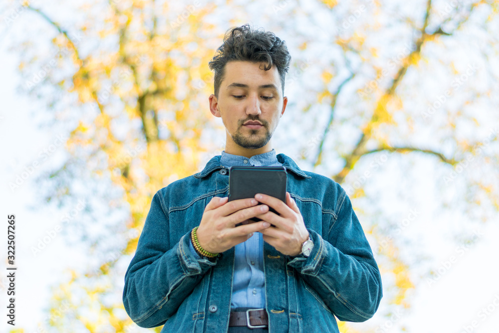 Handsome young man reading an ebook in a park. Portrait of a young man with a denim jacket and blue shirt reading an e-book outside. A guy reading an ebook in a park
