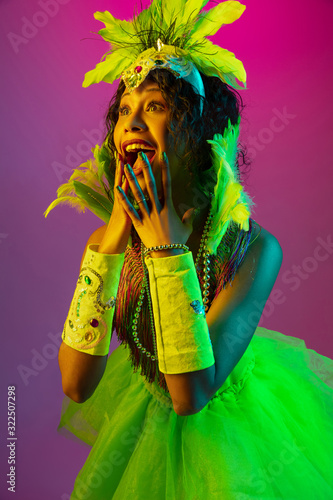 Astonished. Beautiful young woman in carnival, stylish masquerade costume with feathers dancing on gradient background in neon. Concept of holidays celebration, festive time, dance, party, having fun.