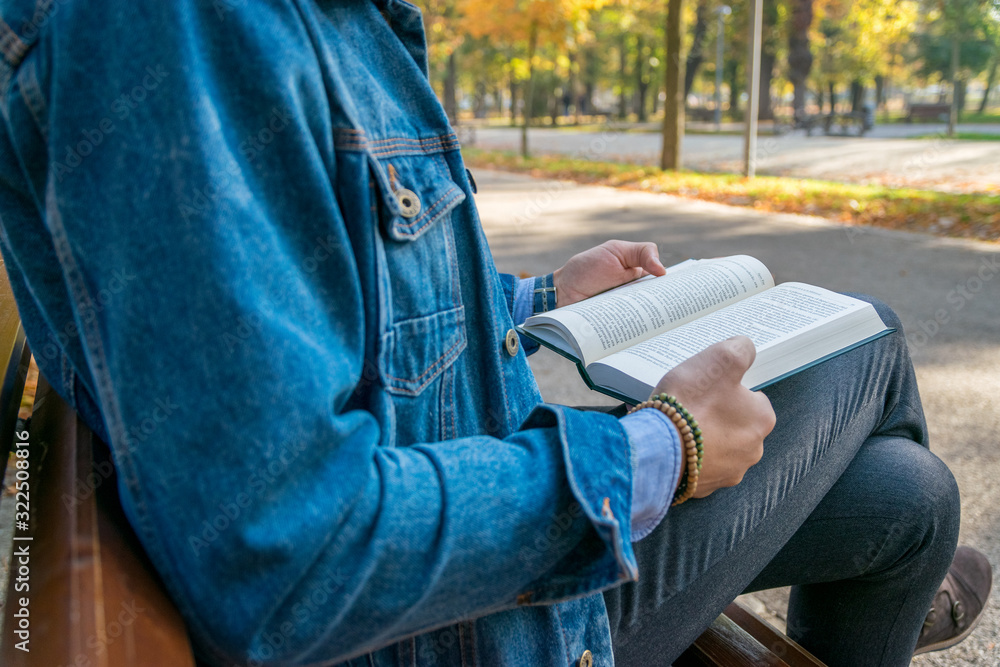 Handsome young man reading a book in a park. Portrait of a young man with a denim jacket and blue shirt reading a book outside. A guy reading a book in a park