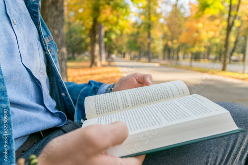 Handsome young man reading a book in a park. Portrait of a young man with a denim jacket and blue shirt reading a book outside. A guy reading a book in a park