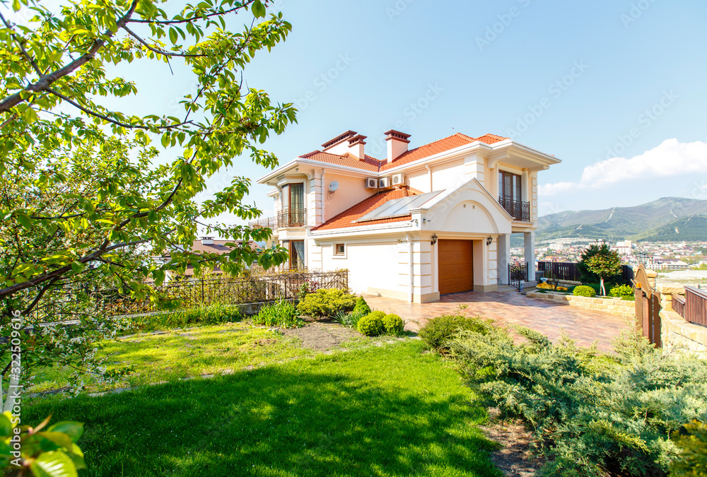 The facade of a classic Mediterranean two-storey cottage in the spring. In front of the cottage there is a green lawn and flowering trees. Mountains in the background. The facade of a classic Mediterranean two-storey cottage in the spring. In front of the cottage there is a green lawn and flowering trees. Mountains in the background.