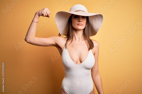 Young beautiful brunette woman on vacation wearing swimsuit and summer hat Strong person showing arm muscle, confident and proud of power