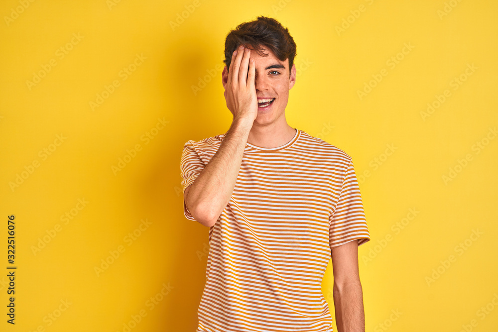 Teenager boy wearing yellow t-shirt over isolated background covering one eye with hand, confident smile on face and surprise emotion. Teenager boy wearing yellow t-shirt over isolated background covering one eye with hand, confident smile on face and surprise emotion.