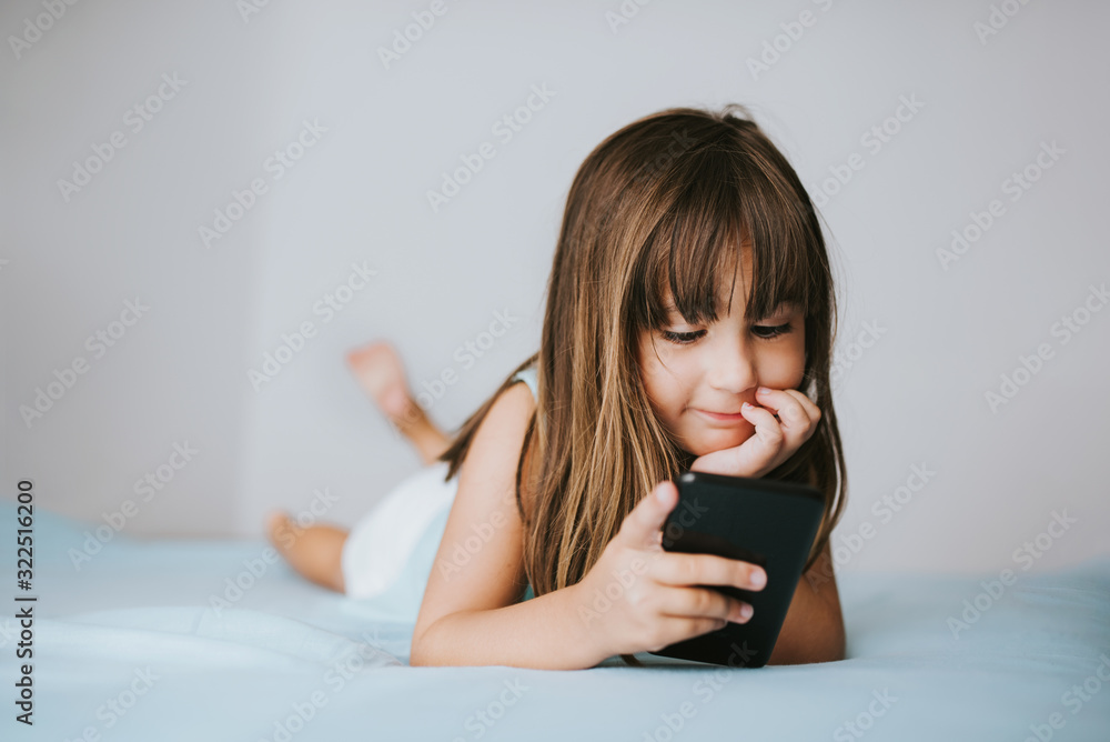 Young girl lying on bed and watching in mobile phone. Children and technology concept. Young girl lying on bed and watching in mobile phone. Children and technology concept.