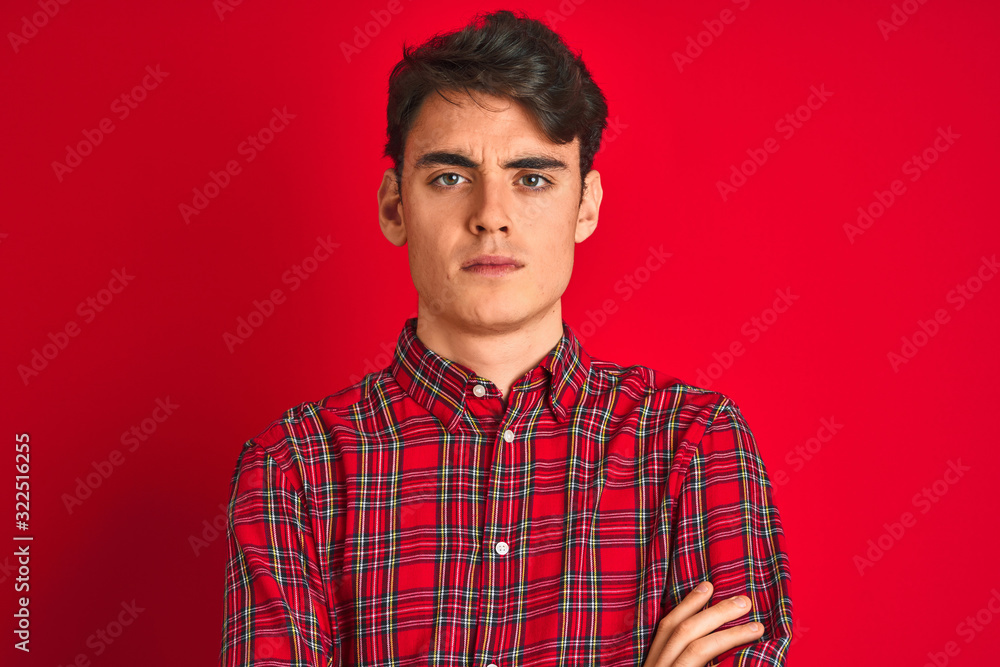 Teenager boy wearing red shirt standing over isolated background skeptic and nervous, disapproving expression on face with crossed arms. Negative person. Teenager boy wearing red shirt standing over isolated background skeptic and nervous, disapproving expression on face with crossed arms. Negative person.