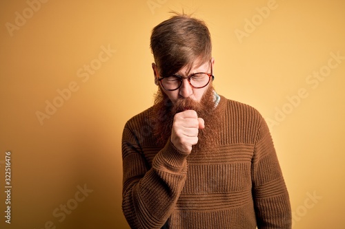 Handsome Irish redhead man with beard wearing glasses and winter sweater over yellow background feeling unwell and coughing as symptom for cold or bronchitis. Health care concept. © Krakenimages.com