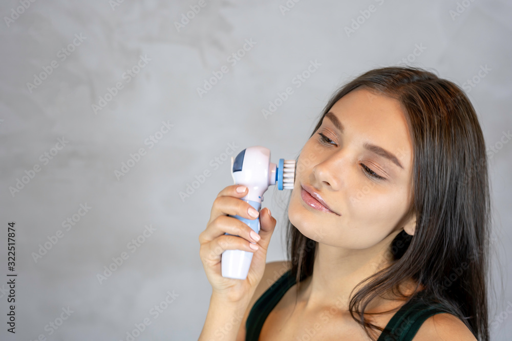 Young pretty woman doing skin care procedure. Cleaning face with machine on grey background. copy space. Young pretty woman doing skin care procedure. Cleaning face with machine on grey background. copy space.