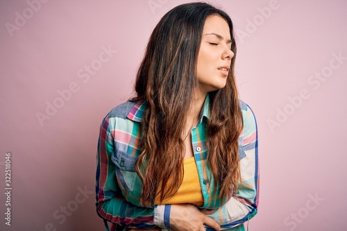 Young beautiful brunette woman wearing casual colorful shirt standing over pink background with hand on stomach because nausea, painful disease feeling unwell. Ache concept.