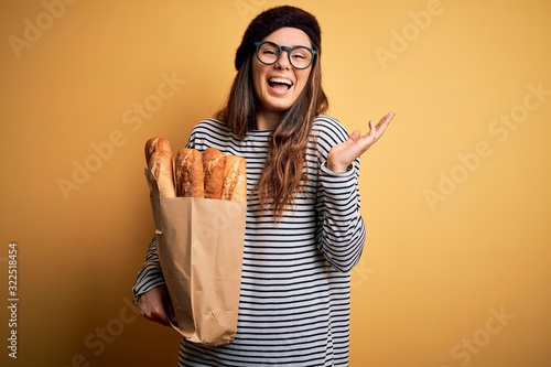 Young beautiful brunette french woman wearing beret holding paper bag with baguettes very happy and excited, winner expression celebrating victory screaming with big smile and raised hands