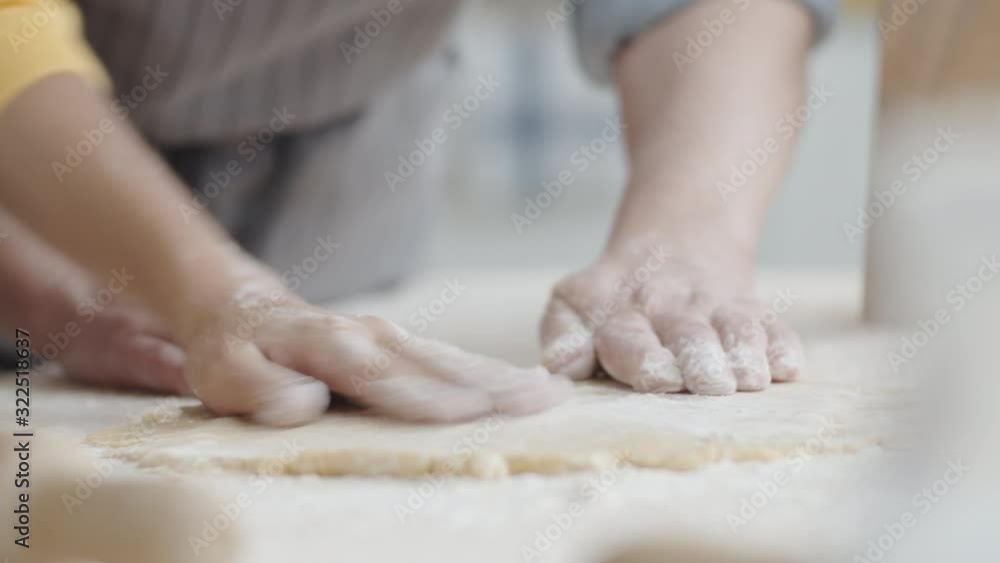 Close up shot of hands of grandmother and little granddaughter sifting flour on pastry dough and rolling it out with wooden pin on table Close up shot of hands of grandmother and little granddaughter sifting flour on pastry dough and rolling it out with wooden pin on table