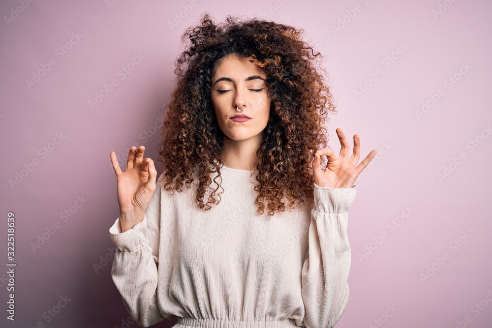 Beautiful woman with curly hair and piercing wearing casual sweater over pink background relax and smiling with eyes closed doing meditation gesture with fingers. Yoga concept. Beautiful woman with curly hair and piercing wearing casual sweater over pink background relax and smiling with eyes closed doing meditation gesture with fingers. Yoga concept.