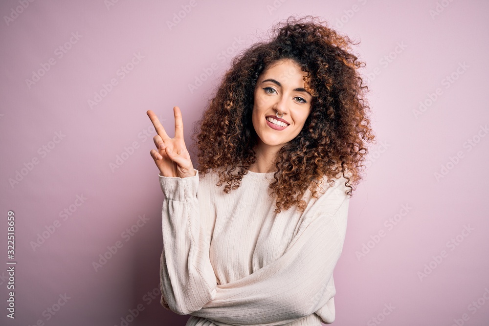 Beautiful woman with curly hair and piercing wearing casual sweater over pink background showing and pointing up with fingers number two while smiling confident and happy. Beautiful woman with curly hair and piercing wearing casual sweater over pink background showing and pointing up with fingers number two while smiling confident and happy.