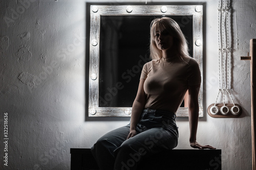 European young fashion model of a beautiful woman sits in a dark room on a table near a vintage white mirror for makeup with bright modern lamps. Sexy blonde girl resting in the bedroom in the evening