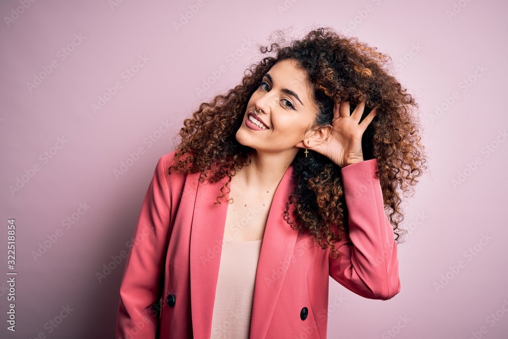 Young beautiful businesswoman with curly hair and piercing wearing elegant jacket smiling with hand over ear listening an hearing to rumor or gossip. Deafness concept.