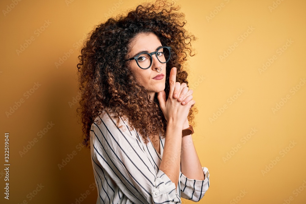 Young beautiful woman with curly hair and piercing wearing striped shirt and glasses Holding symbolic gun with hand gesture, playing killing shooting weapons, angry face Young beautiful woman with curly hair and piercing wearing striped shirt and glasses Holding symbolic gun with hand gesture, playing killing shooting weapons, angry face