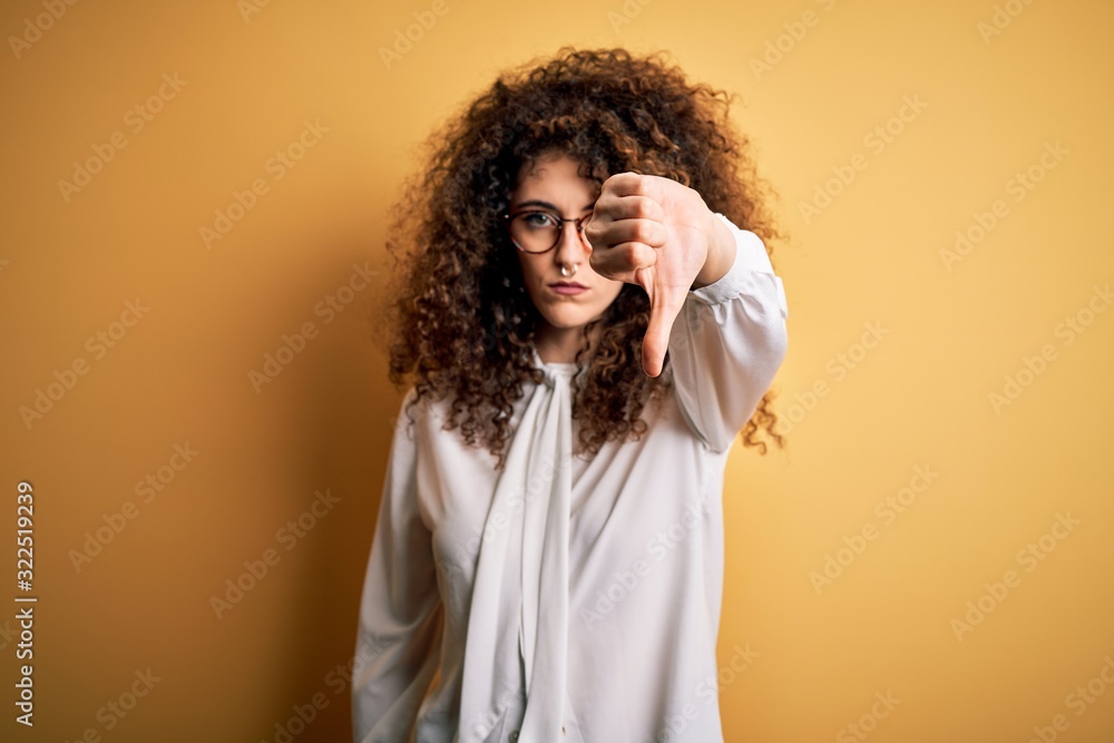 Young beautiful brunette woman with curly hair and piercing wearing shirt and glasses looking unhappy and angry showing rejection and negative with thumbs down gesture. Bad expression. Young beautiful brunette woman with curly hair and piercing wearing shirt and glasses looking unhappy and angry showing rejection and negative with thumbs down gesture. Bad expression.
