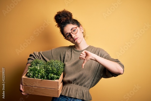 Young beautiful gardener woman with curly hair and piercing holding wooden box with plants with angry face, negative sign showing dislike with thumbs down, rejection concept