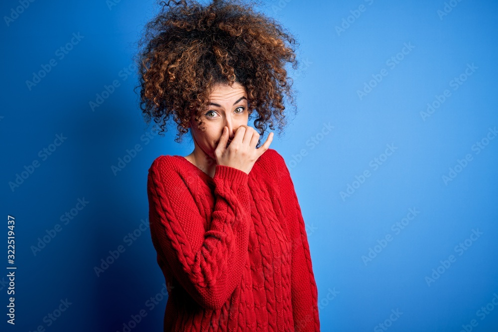 Young beautiful woman with curly hair and piercing wearing casual red sweater smelling something stinky and disgusting, intolerable smell, holding breath with fingers on nose. Bad smell Young beautiful woman with curly hair and piercing wearing casual red sweater smelling something stinky and disgusting, intolerable smell, holding breath with fingers on nose. Bad smell