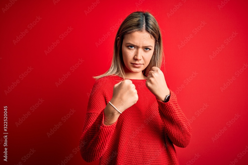 Young beautiful blonde woman wearing casual sweater over red isolated background Ready to fight with fist defense gesture, angry and upset face, afraid of problem Young beautiful blonde woman wearing casual sweater over red isolated background Ready to fight with fist defense gesture, angry and upset face, afraid of problem