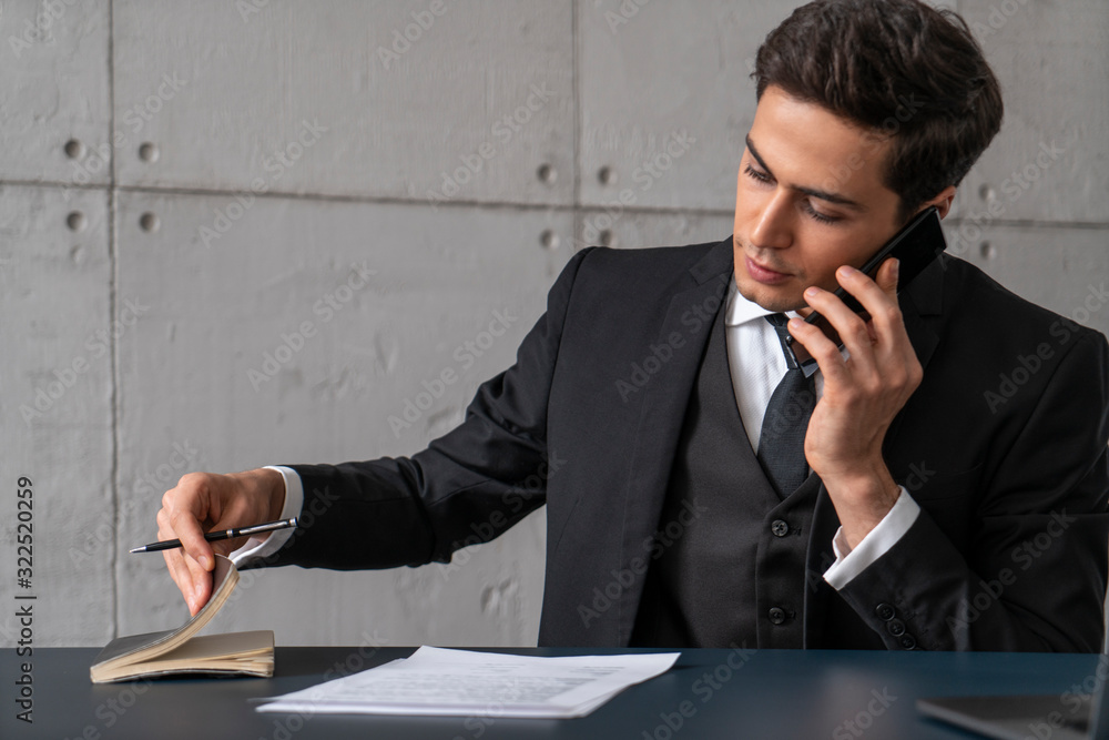 Serious young businessman in dark suit talking on smartphone and checking his schedule at loft office table with documents. Concept of paperwork Serious young businessman in dark suit talking on smartphone and checking his schedule at loft office table with documents. Concept of paperwork
