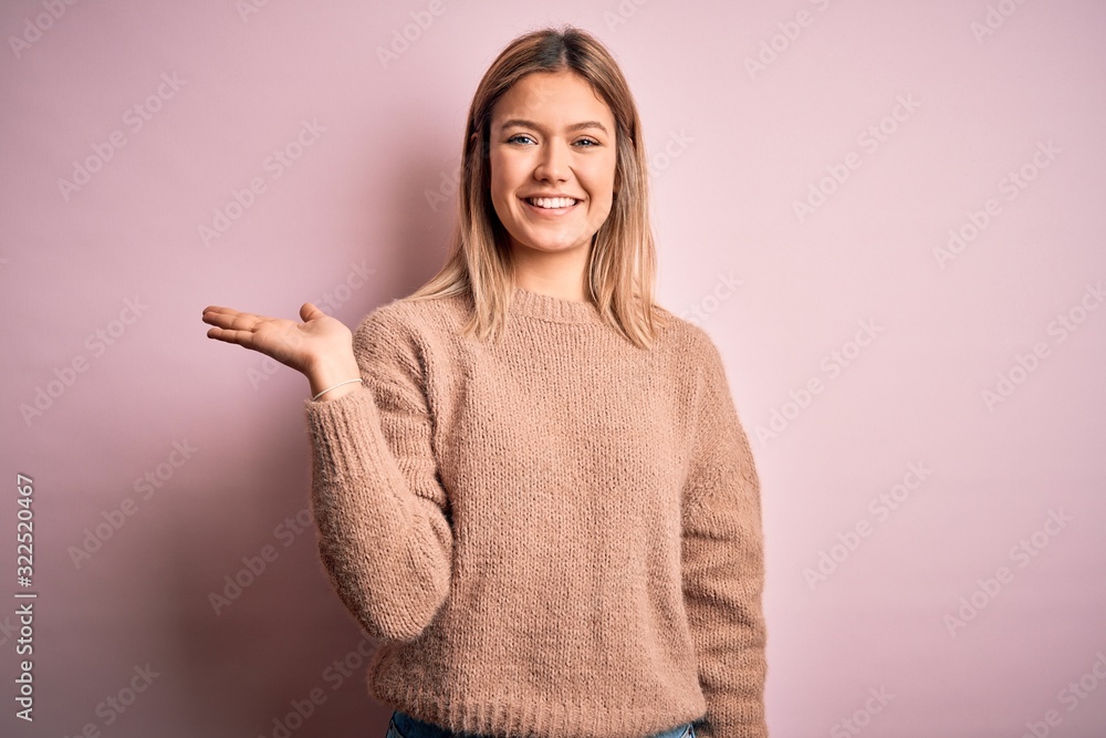 Young beautiful blonde woman wearing winter wool sweater over pink isolated background smiling cheerful presenting and pointing with palm of hand looking at the camera. Young beautiful blonde woman wearing winter wool sweater over pink isolated background smiling cheerful presenting and pointing with palm of hand looking at the camera.