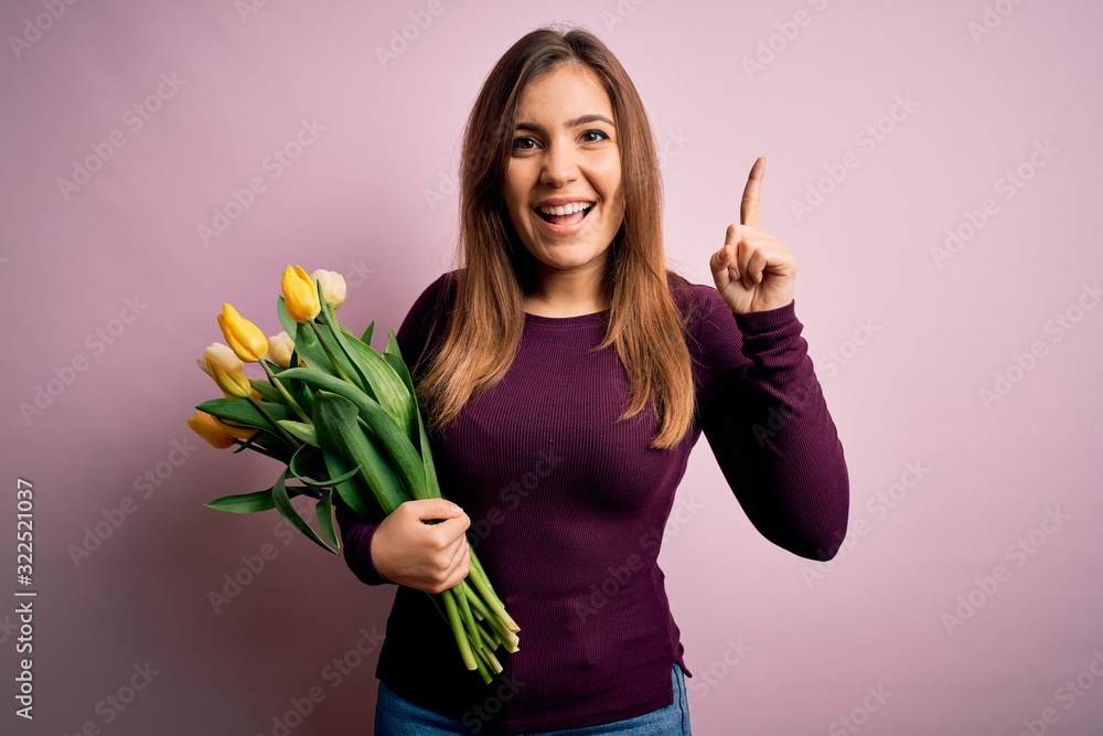 Young blonde woman holding romantic bouquet of yellow tulips flowers over pink background smiling amazed and surprised and pointing up with fingers and raised arms. Young blonde woman holding romantic bouquet of yellow tulips flowers over pink background smiling amazed and surprised and pointing up with fingers and raised arms.