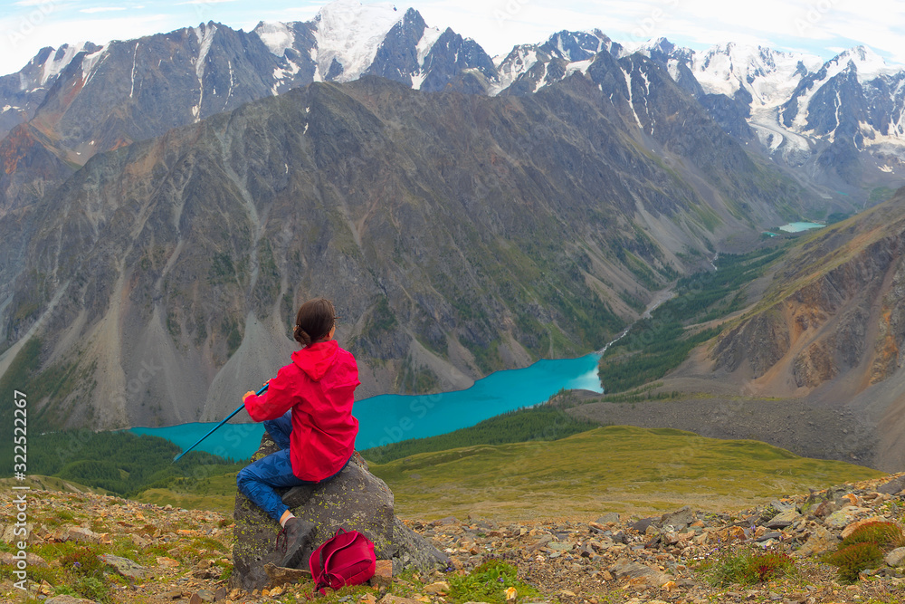 Fototapeta premium A girl in a red jacket is sitting on a high mountain. A woman is looking at a mountain lake. A tourist admires the panorama of the mountains.