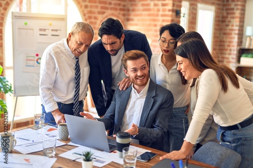 Group of business workers smiling happy and confident. One of them sitting and partners standing around. Working together with smile on face looking at the laptop at the office