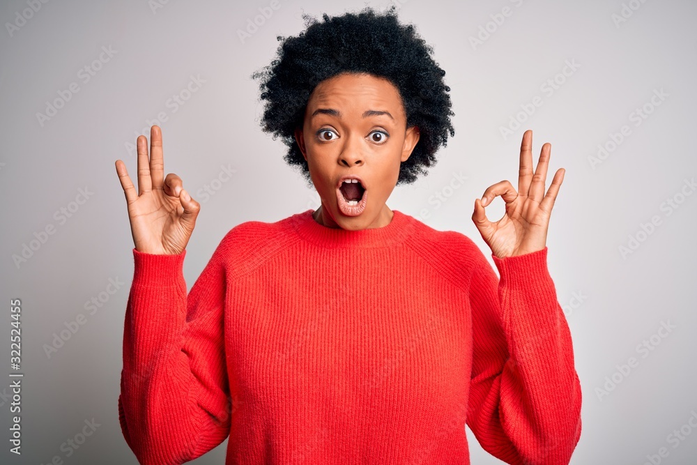 Young beautiful African American afro woman with curly hair wearing red casual sweater looking surprised and shocked doing ok approval symbol with fingers. Crazy expression Young beautiful African American afro woman with curly hair wearing red casual sweater looking surprised and shocked doing ok approval symbol with fingers. Crazy expression
