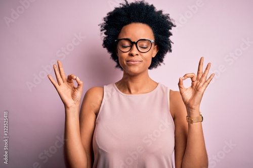 Young beautiful African American afro woman with curly hair wearing t-shirt and glasses relax and smiling with eyes closed doing meditation gesture with fingers. Yoga concept.