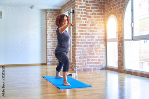 Fototapeta Naklejka Na Ścianę i Meble -  Middle age beautiful sportwoman smiling happy. Standing on mat practicing yoga doing warrior pose at gym