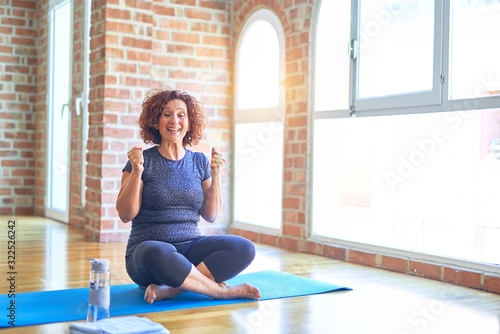 Middle age beautiful sportswoman wearing sportswear sitting on mat practicing yoga at home celebrating surprised and amazed for success with arms raised and open eyes. Winner concept.