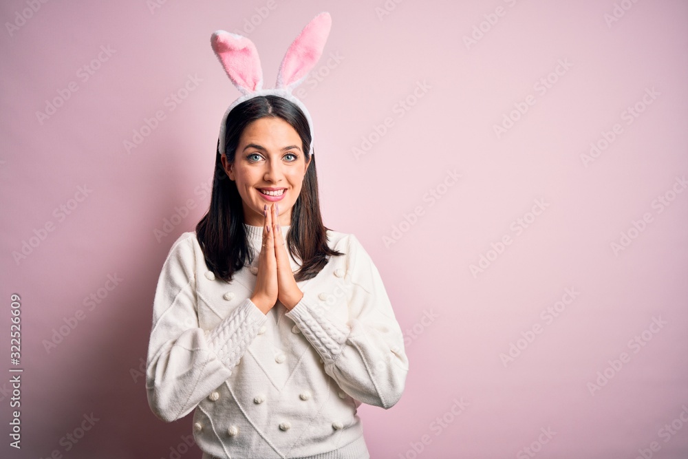 Young caucasian woman wearing cute easter rabbit ears over pink isolated background praying with hands together asking for forgiveness smiling confident.