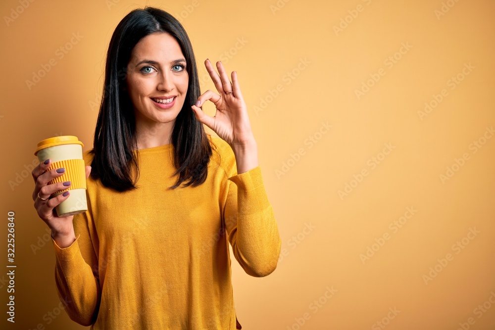 Young woman with blue eyes holding cup of coffee standing over yellow background doing ok sign with fingers, excellent symbol Young woman with blue eyes holding cup of coffee standing over yellow background doing ok sign with fingers, excellent symbol